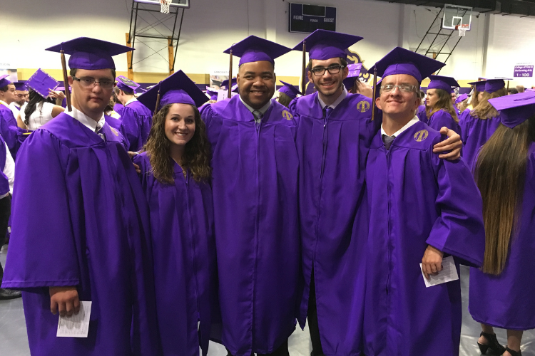 Photo of a group of five students smiling at a college graduation, each wearing a purple cap and gown.