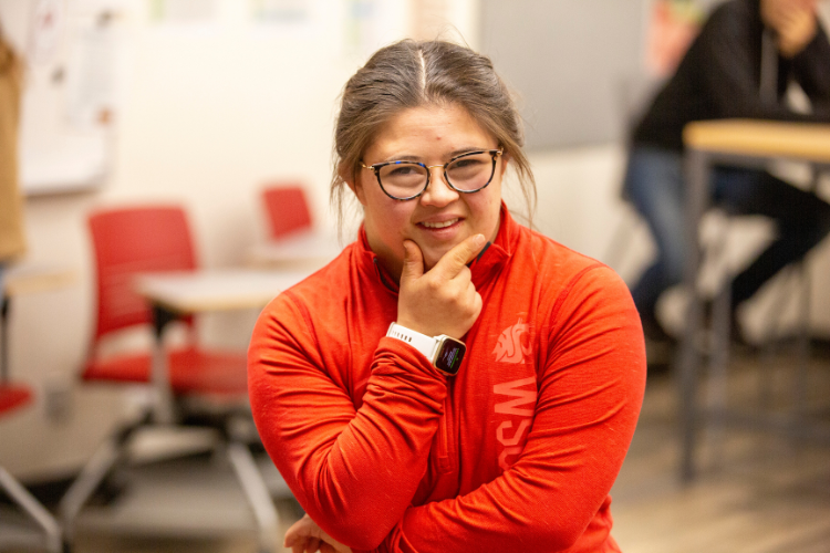 A female student with brown hair and a red sweatshirt smiles looking at the camera resting her hand on her chin.