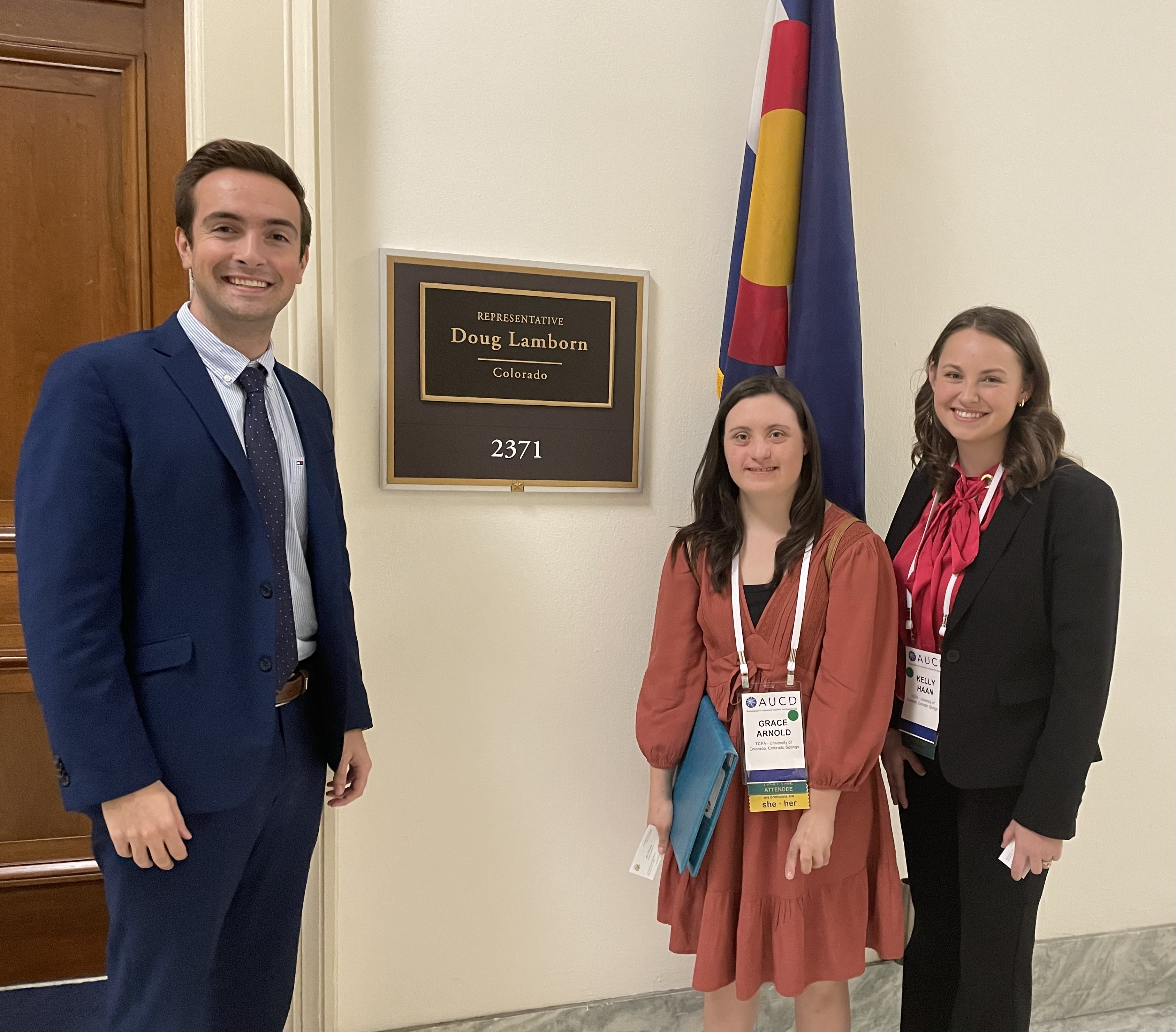 Two women standing with a man in front of Representative Doug Lamborn's office. The Colorado state flag is behind them. 