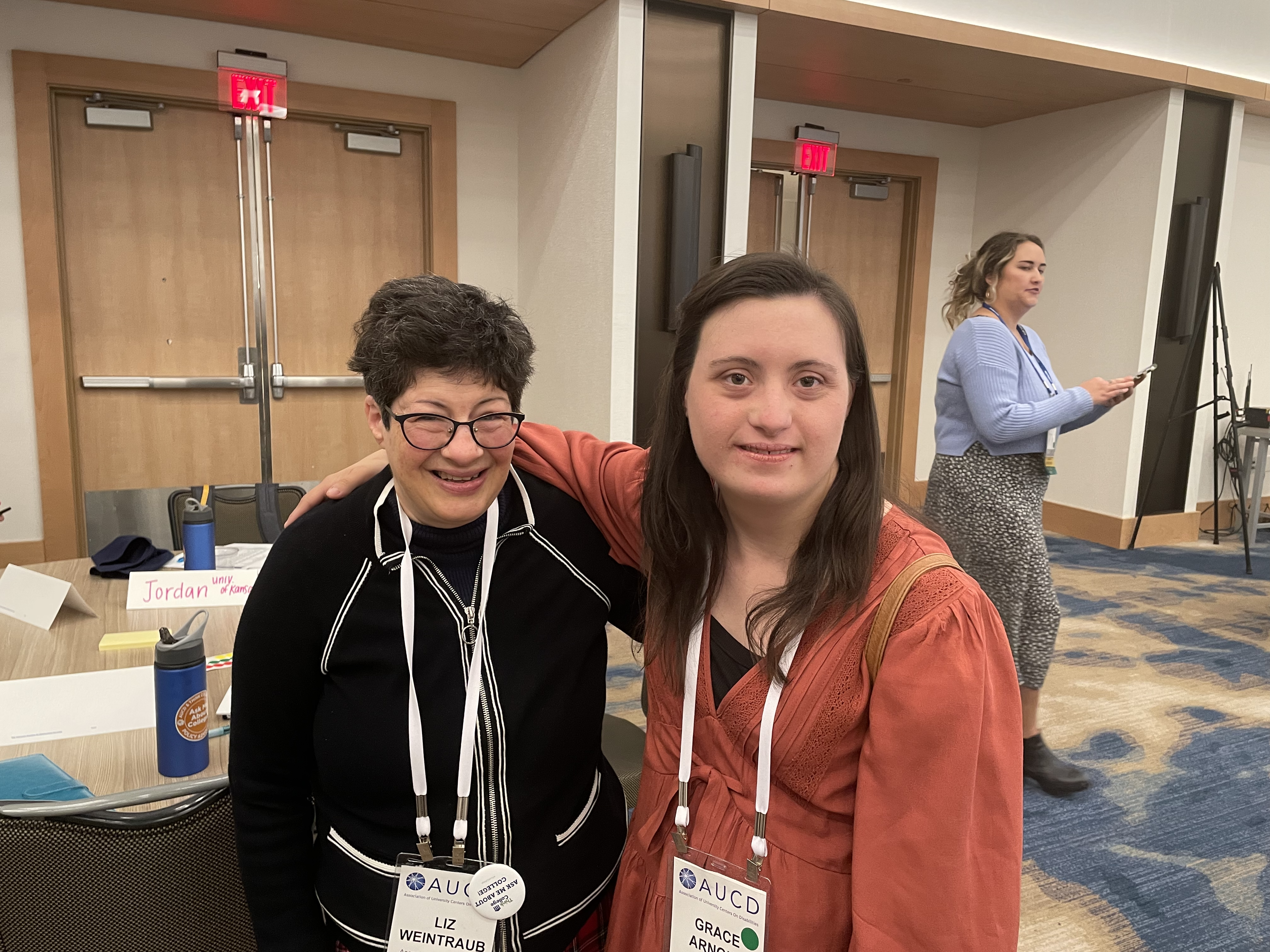 Two women with arms around each other, smiling at the camera. The one on the left has short curly dark brown hair and glasses, the one on the right has shoulder length straight brown hair.