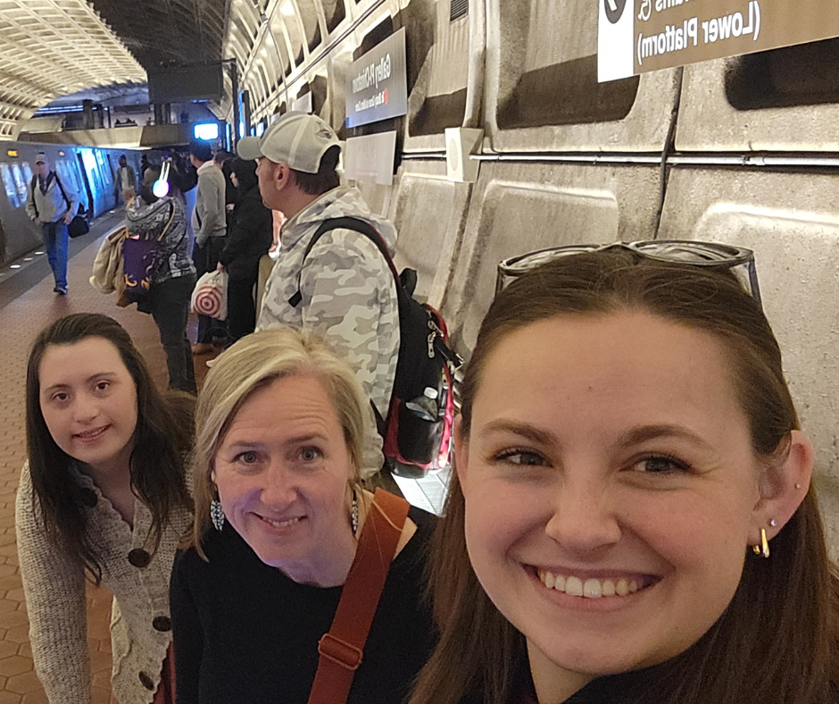 Three white women, two with shoulder length straight brown hair, the one in the middle with straight blond hair, all smiling looking at the camera, while standing in an underground train station.