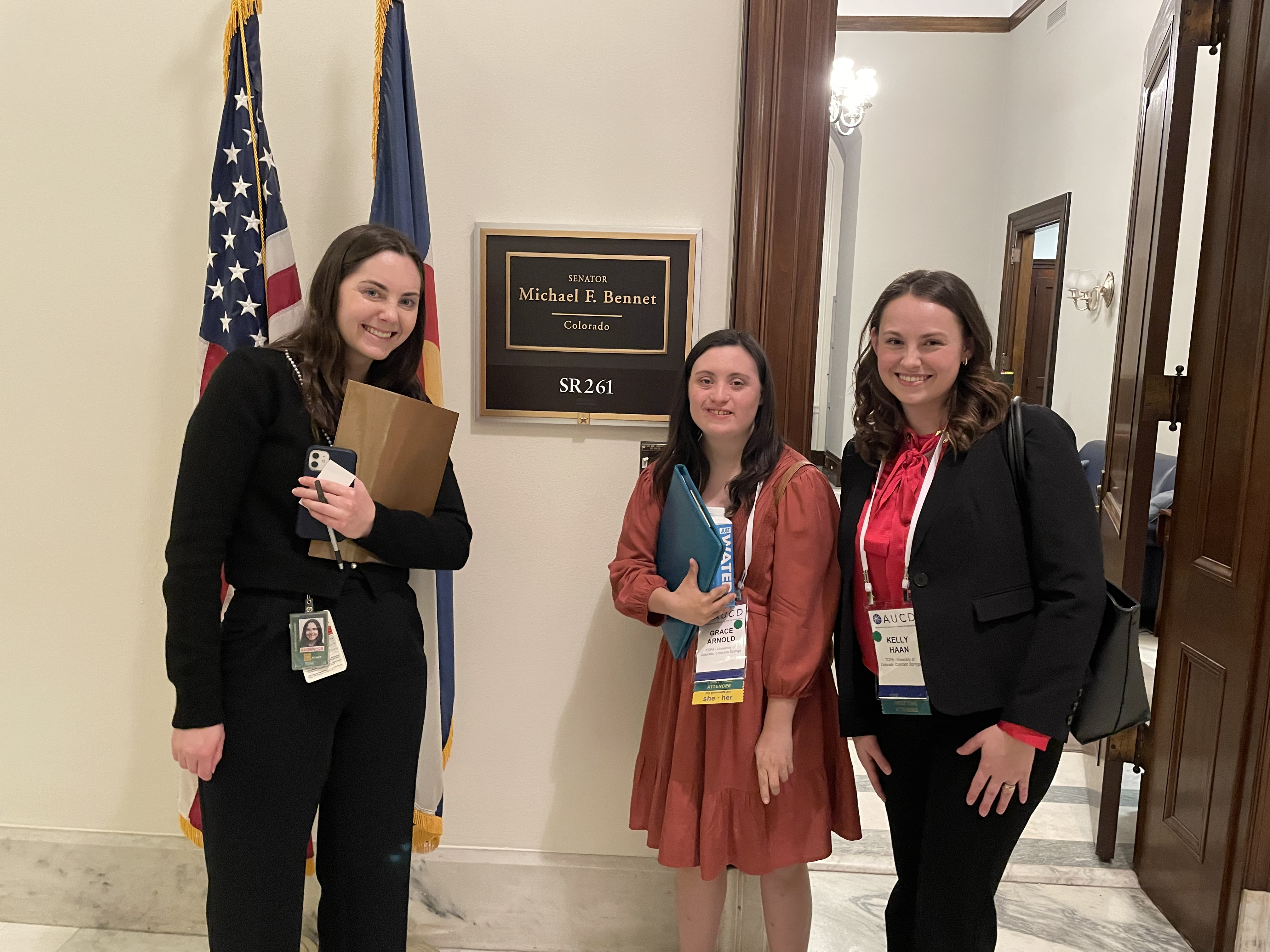 Three women standing in front of Sen. Michael F. Bennet's office door. The US and Colorado state flag are behind them