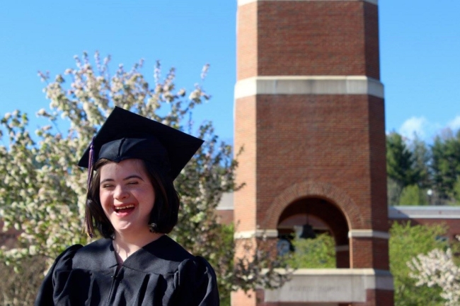 Woman with disability stands in cap and gown in front of campus clock tower building