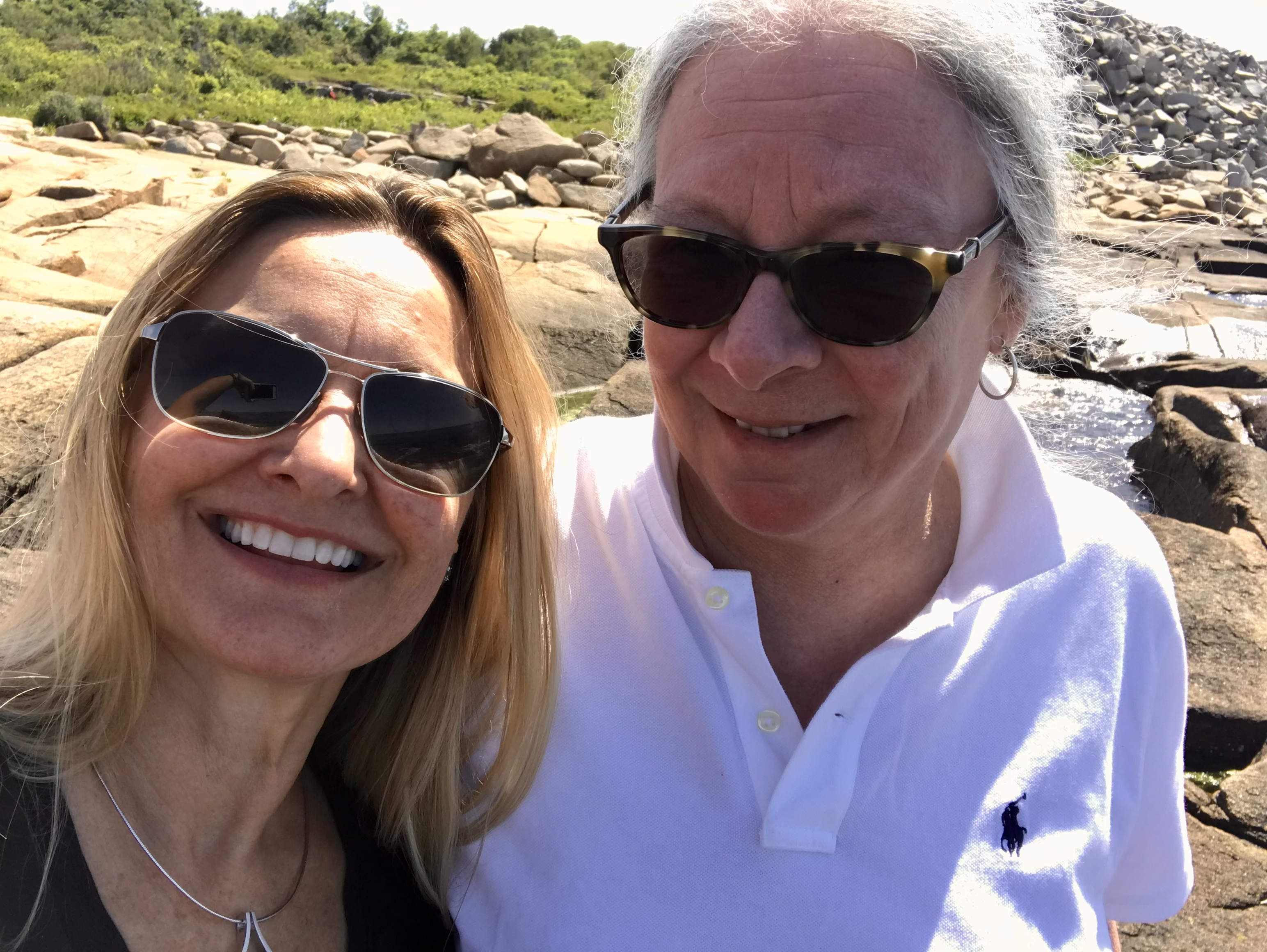 Meg Grigal and Debra Hart standing together smiling, wearing sunglasses, at a rocky beach in Rhode Island.