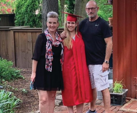 Young woman in red cap and gown stands on outdoor patio with mom and bonus dad