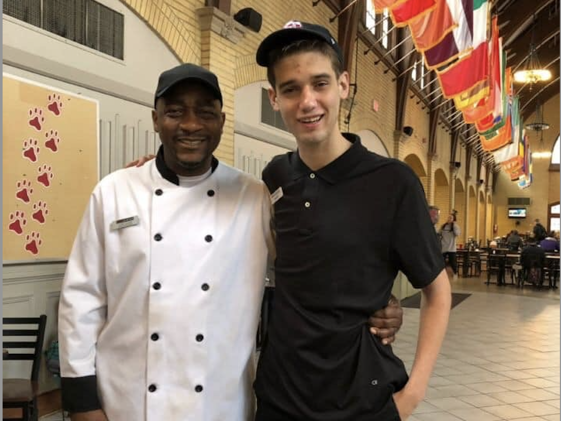 Image of two men with arms around each other, one in a culinary jacket and one in black shirt and baseball cap