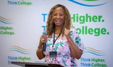 African American woman with medium length brown hair in a pink floral dress speaking into a microphone in front of a banner that says “Think Higher. Think College.”