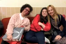 Photo of 3 women sitting in a hallway on college campus, smiling. Left to right: woman in pink sweater, short dark curly hair and glasses, holding a binder; woman in black shirt with red sweater, arms and legs crossed; woman with long blond hair, leaning on woman in red sweater