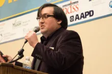 photo of Kenneth Kelty speaking at a conference.  he is holding a microphone and standing behind a lectern.