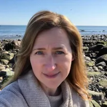 image of a woman smiling with dark blond hair standing on a rocky shoreline in front of the ocean
