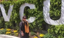 student in graduation gown standing in front of the campus entrance sign