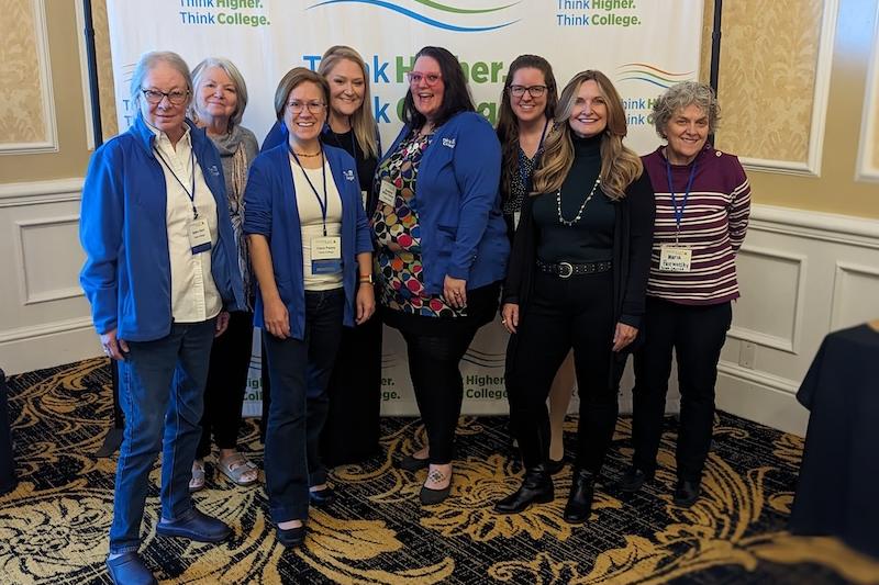 Group of women, several wearing blue Think College sweaters, standing in front of a Think Higher. Think College. backdrop