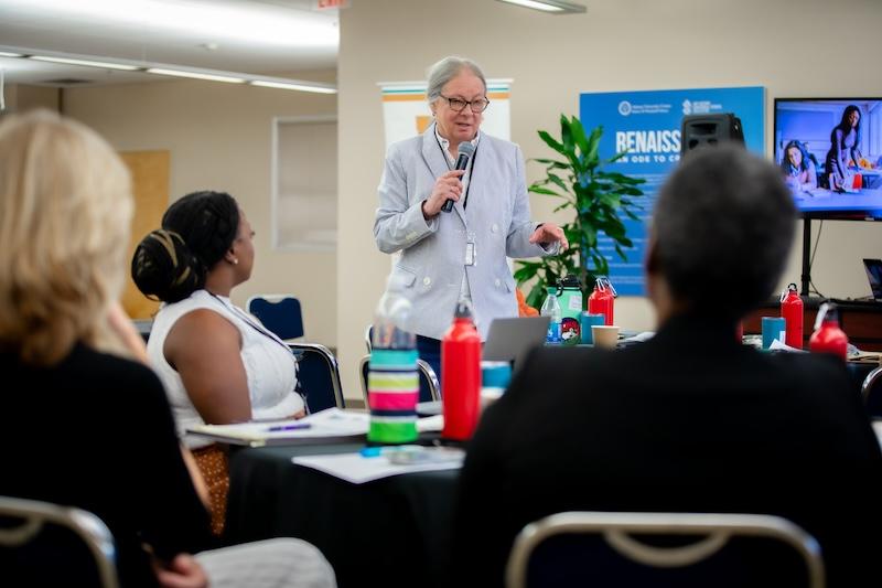 White woman with grey hair, talking into a microphone, standing in front of some people seated at tables