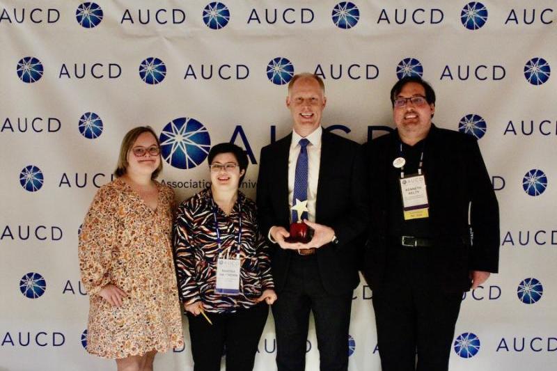Four people standing together smiling, two college students and a college alumni, with the Senior Education Policy Advisor.