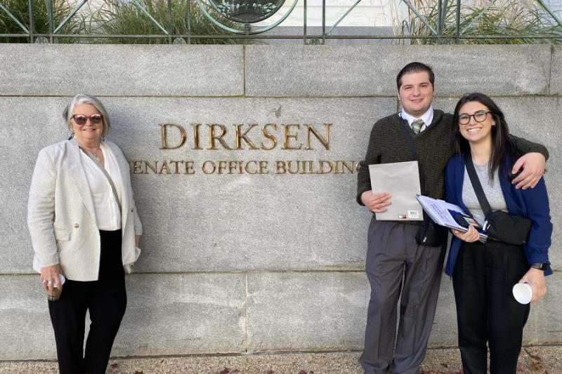 Three people smiling, standing in front of the Dirksen Senate Office Building sign.