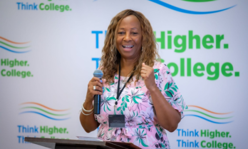 African American woman with medium length brown hair in a pink floral dress speaking into a microphone in front of a banner that says “Think Higher. Think College.”